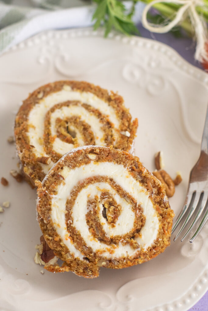 Carrot Cake Roll Slices on an off-white plate next to a fork with fresh carrots in the background.