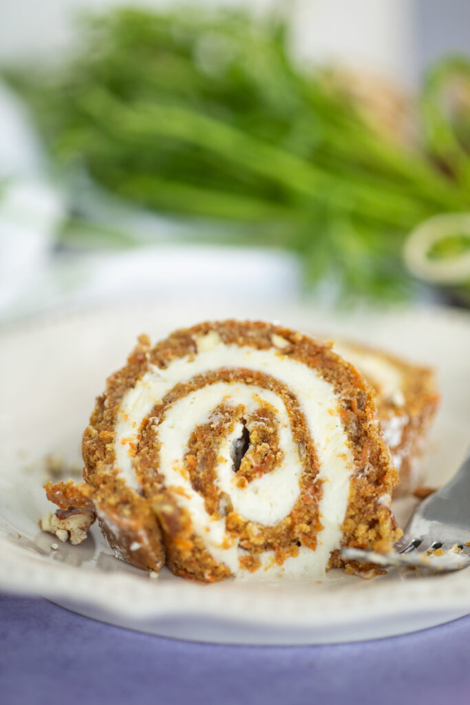 Side view of a carrot cake roll slice with cream cheese swirls with a bite taken out with fresh carrots in the background.