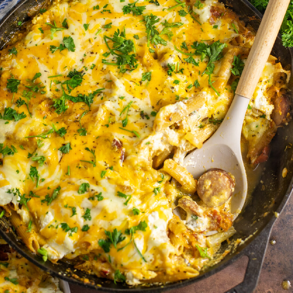 Cheesy baked kielbasa pasta in a cast iron skillet topped with freshly chopped parsley with a wooden spoon holding a scoop of the pasta.