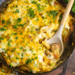 Cheesy baked kielbasa pasta in a cast iron skillet topped with freshly chopped parsley with a wooden spoon holding a scoop of the pasta.