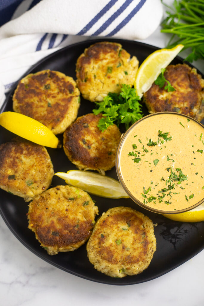 Overhead view of lump crab cakes on a large matte black plate with lemon wedges, a sprig of fresh parsley, and a small glass bowl filled with homemade remoulade for dipping.