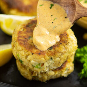A wooden spoon drizzling homemade remoulade on top of a lump crab cake on a black plate next to lemon wedges and fresh parsley.