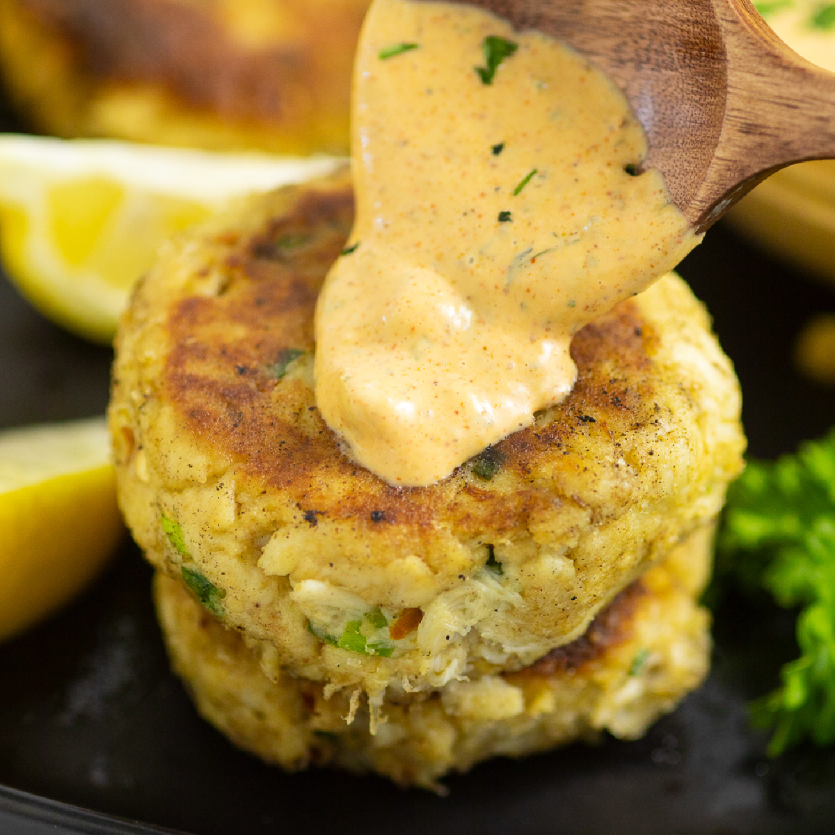 A wooden spoon drizzling homemade remoulade on top of a lump crab cake on a black plate next to lemon wedges and fresh parsley.
