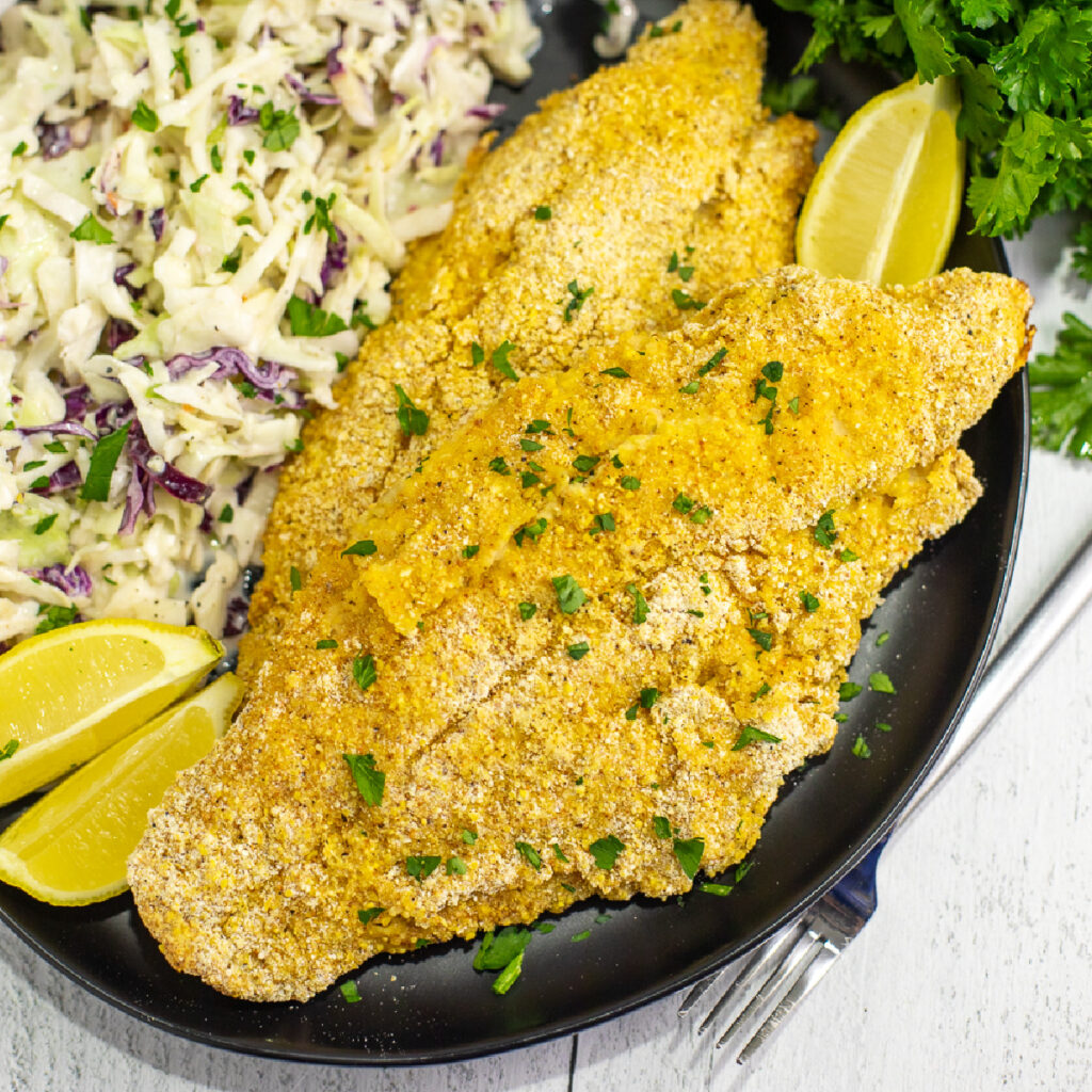 Two oven fried catfish filets on a black plate next to coleslaw and lemon wedges, sprinkled with freshly chopped parsley with a bunch of fresh parsley above it.