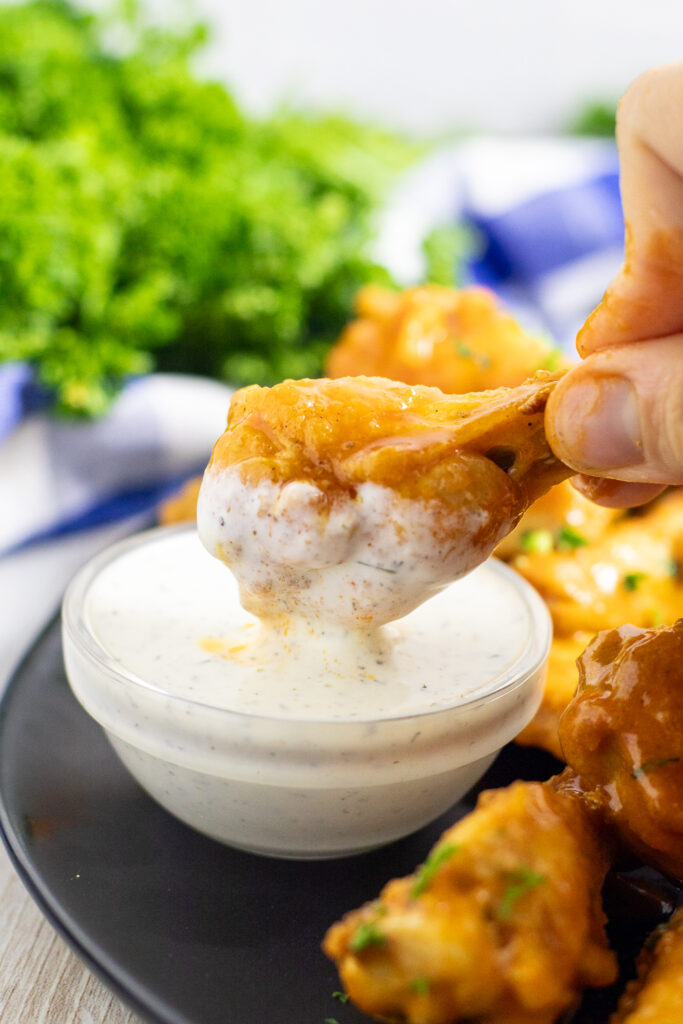 A buffalo chicken wing dipping into a small glass bowl of homemade ranch surrounded by more chicken wings with a bunch of fresh parsley behind.