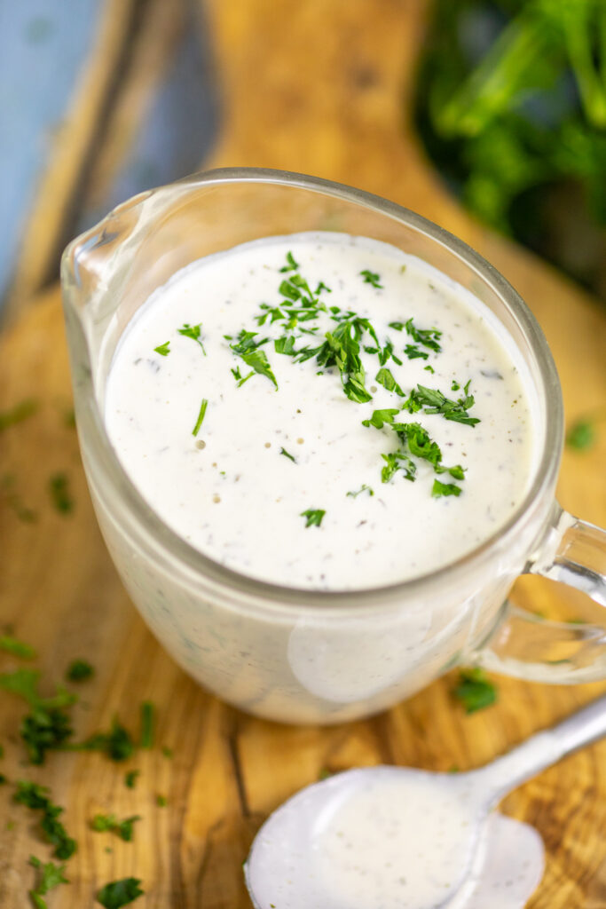 Overhead view of homemade ranch in a small glass pitcher topped with chopped parsley on a small wooden cutting board.