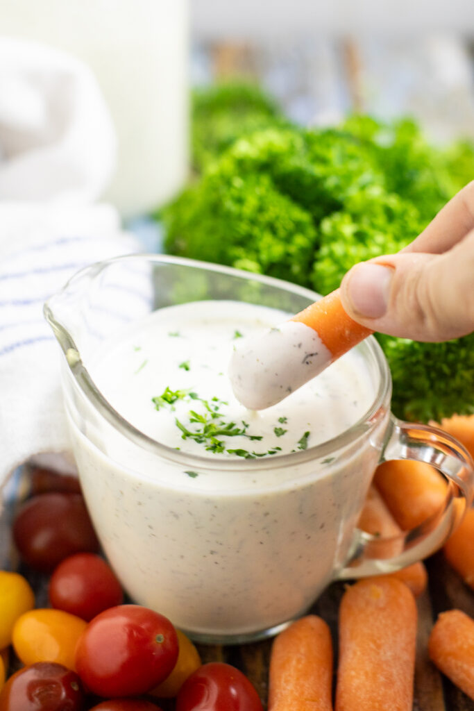 A baby carrot dipping into homemade ranch in a small glass pitcher surrounded by cherry tomatoes, more baby carrots and a bunch of parsley behind.