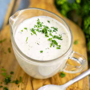 Homemade ranch in a small glass pitcher on a wood cutting board topped with freshly chopped parsley next to a metal spoon.