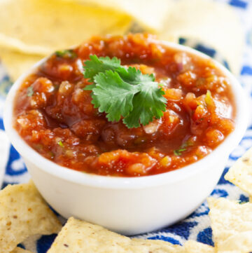 Fresh homemade salsa in a bowl topped with fresh cilantro surrounded by tortilla chips sitting on a colorful linen