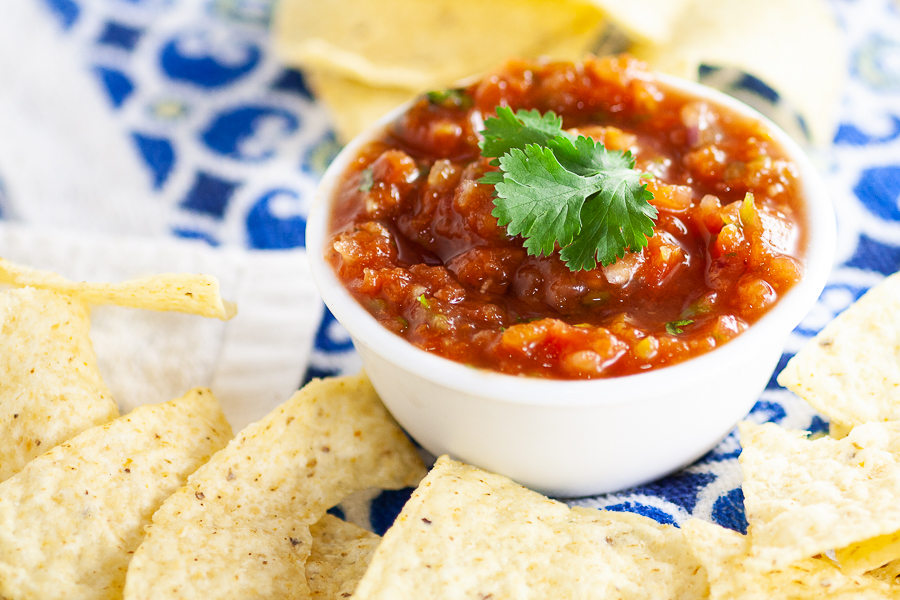 Horizontal image fresh homemade salsa in a bowl topped with fresh cilantro surrounded by tortilla chips sitting on a colorful linen