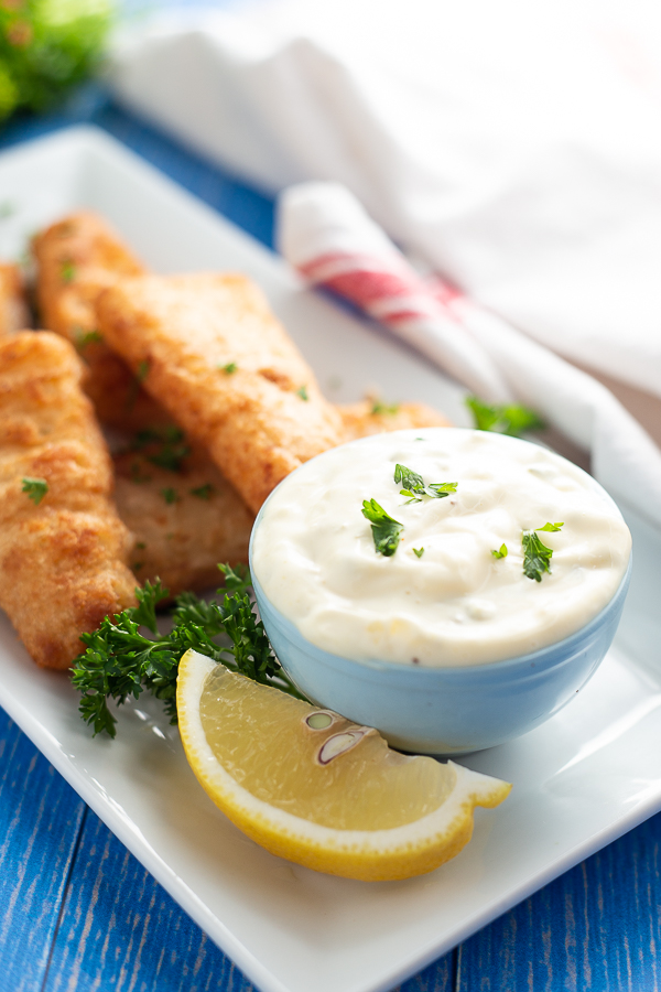 Homemade Tartar sauce in a light blue bowl with a lemon wedge in front and fish behind.