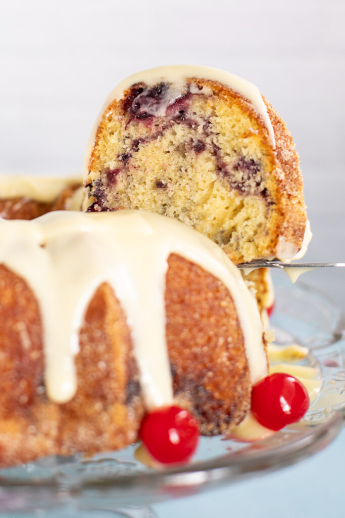 A slice of cherry marble cake being lifted from the full bund cake with cream cheese glaze dripping down the cake on a glass plate with maraschino cherries.