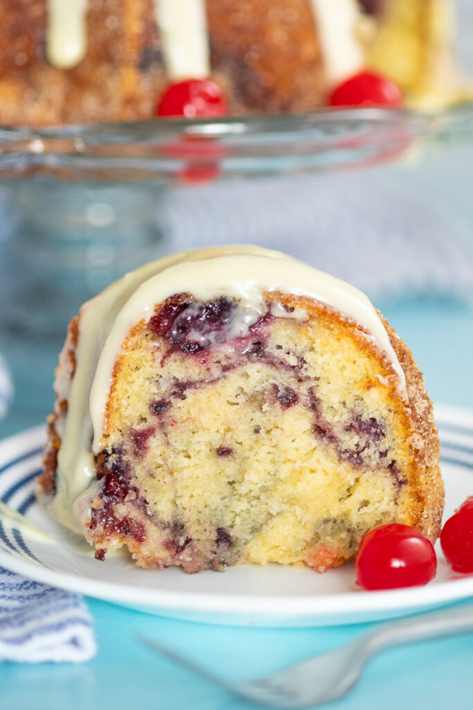 A slice of cherry marble cake topped with cream cheese glaze with a pretty cherry swirl in the center of the slice, sitting on a small white plate next to two maraschino cherries on a bright aqua-blue background.