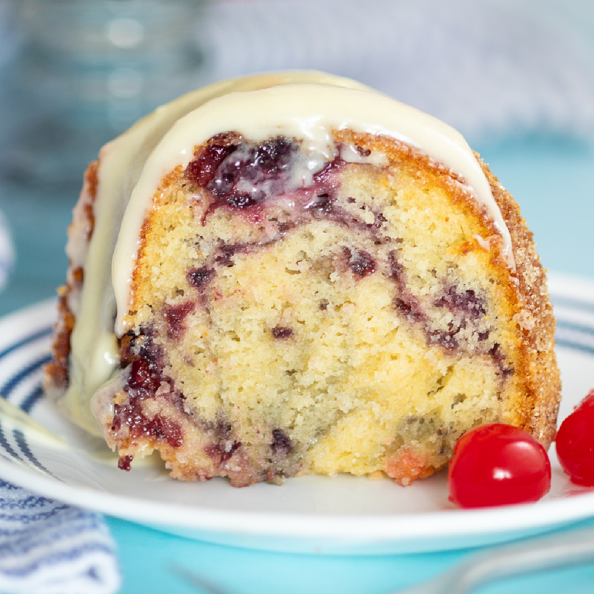 A slice of cherry marble cake topped with cream cheese glaze with a pretty cherry swirl in the center of the slice, sitting on a small white plate next to two maraschino cherries on a bright aqua-blue background.
