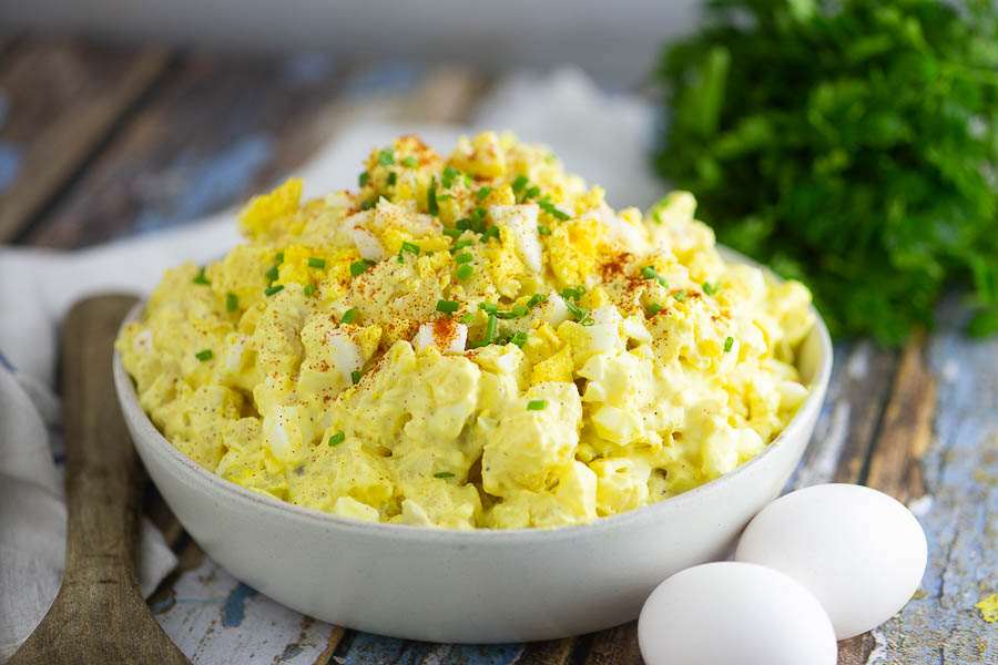 Side shot of deviled egg potato salad in a bowl next to a wooden spoon, 2 eggs, and a bundle of herbs on a rustic wood background