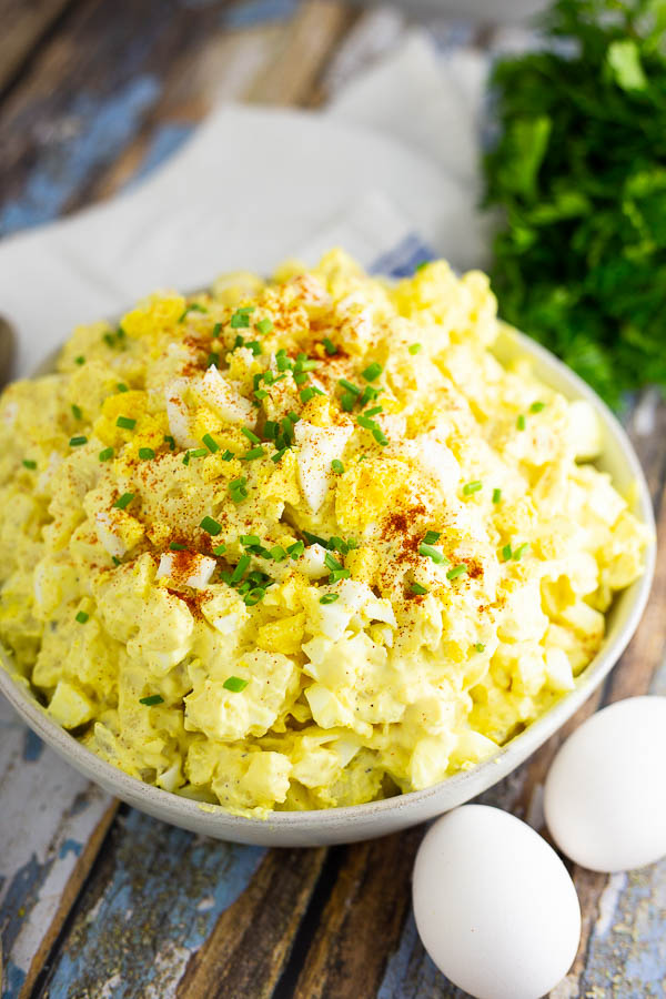 Angle shot of deviled egg potato salad in a wide bowl topped with chives and paprika, next to 2 eggs and herbs on a rustic wooden background