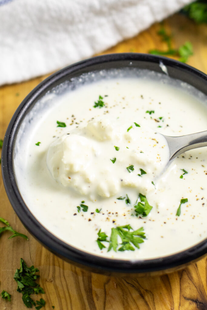 Homemade blue cheese dressing in a small black bowl with a spoon scooping some out in the middle. The bowl is on a wooden cutting board next to a linen napkin.