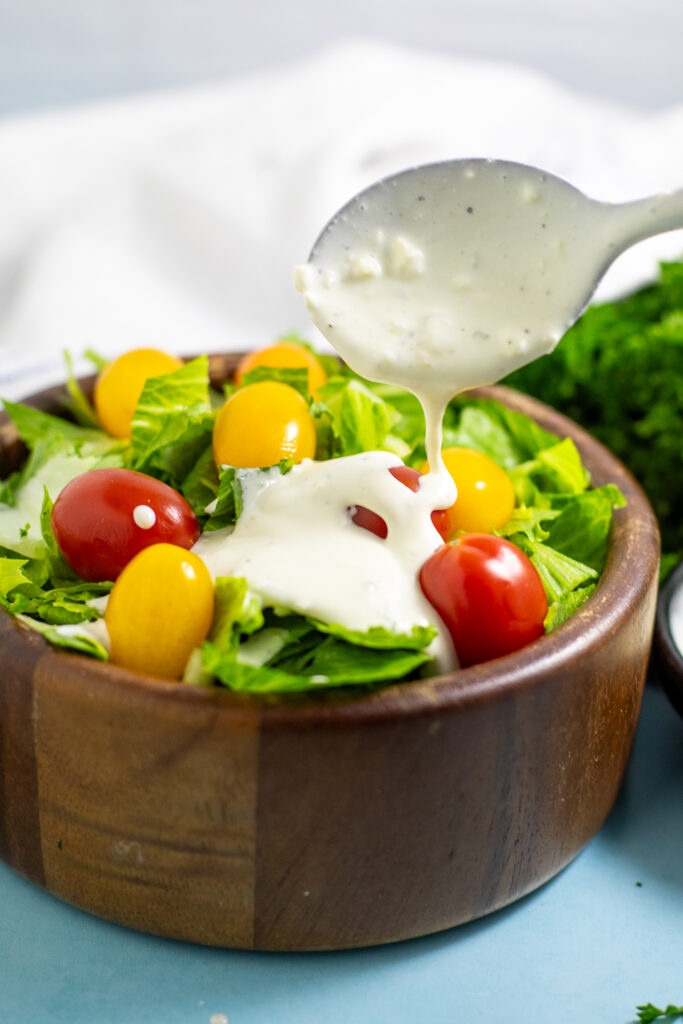 Homemade Blue cheese dressing being drizzled with a spoon onto a green salad with yellow and red cherry tomatoes in a wooden bowl on a bright blue background.