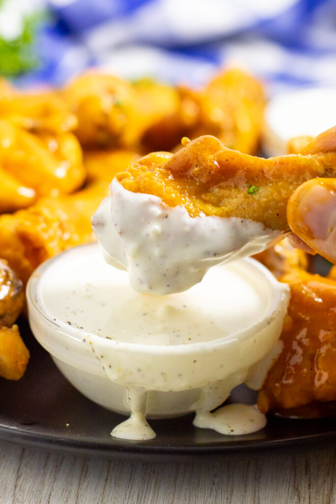A buffalo chicken wing being dipped into a small glass bowl of homemade blue cheese dressing with more wings and a blue checkered linen behind.
