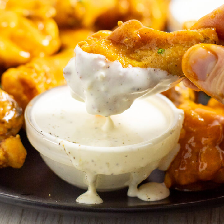 A buffalo chicken wing dipping into a small glass bowl of homemade blue cheese dressing with more wings behind.