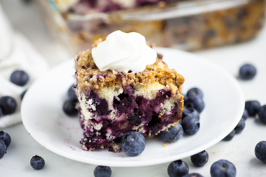 Horizontal image of blueberry buckle topped with whipped cream on a small plate with fresh blueberries scattered around it.