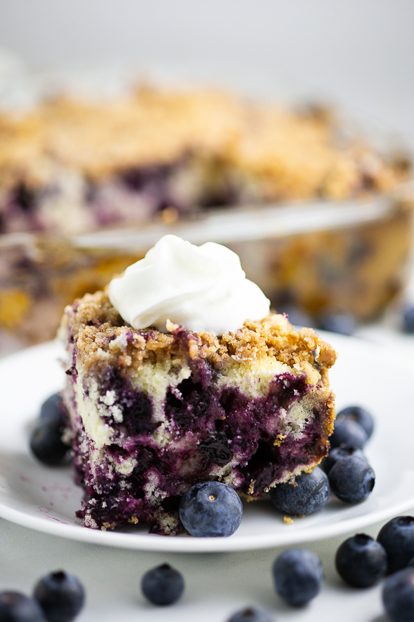 Side view of blueberry buckle topped with whipped cream on a small plate with fresh blueberries with more fresh blueberries in the foreground, and the rest of the cake in the dish behind