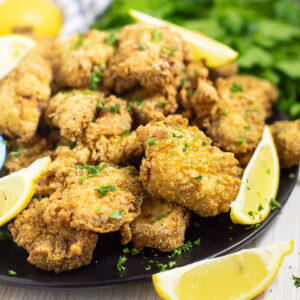 A plate stacked with fried catfish nuggets next to lemon wedges with a bunch of fresh parsley behind.