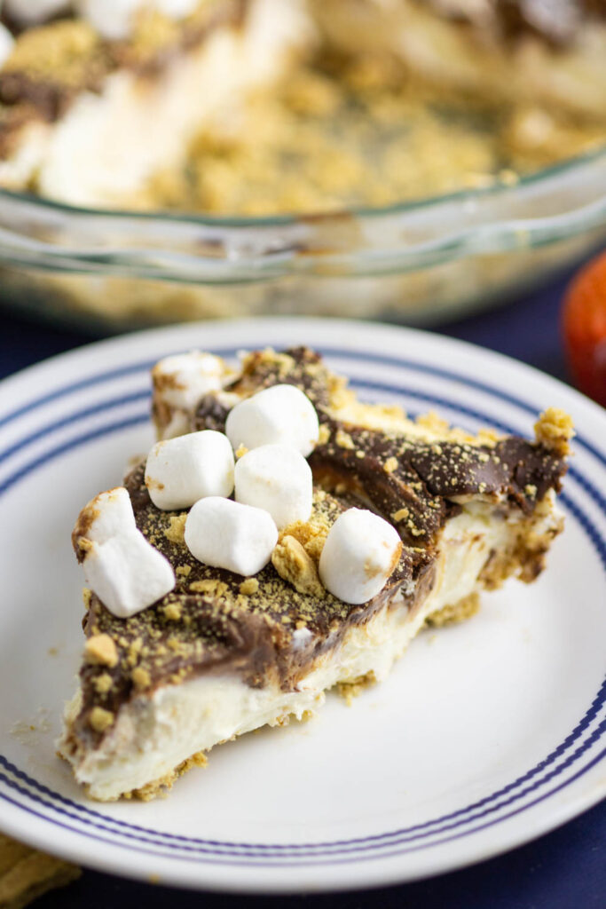 A slice of no bake s'mores cheesecake topped with mini marshmallows and graham cracker crumbs on a small plate with a pie dish behind.