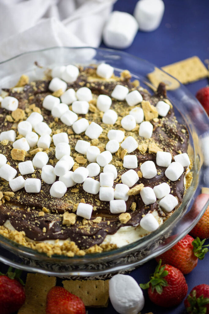 A no bake s'mores cheesecake in a glass pie dish topped with mini marshmallows and graham cracker crumbs, and surrounded by large marshmallows and fresh strawberries.