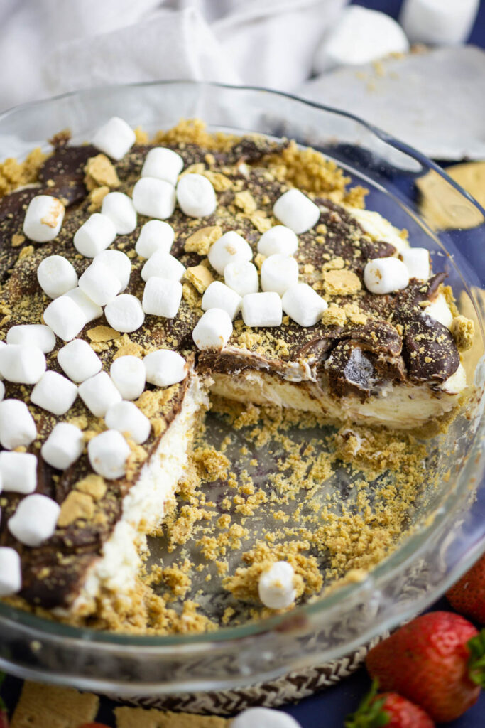 A no bake s'mores cheesecake topped with mini marshmallows and graham cracker crumbs in a glass pie dish with pieces cut out to expose the creamy interior.