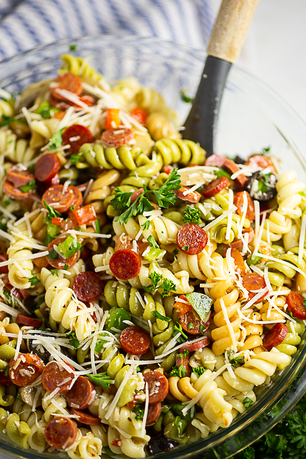 Close up of a glass bowl of pizza pasta salad topped with chopped parsley and grated parmesan with a wooden spoon in it.