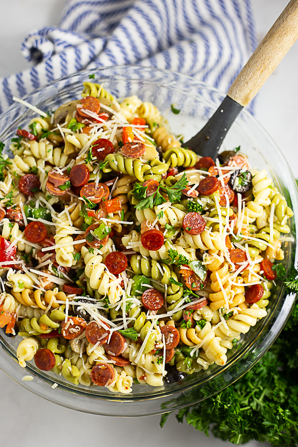 Overhead view of a large bowl of pizza pasta salad with a wooden spoon in it and a striped linen and a bunch of fresh parsley in the background.