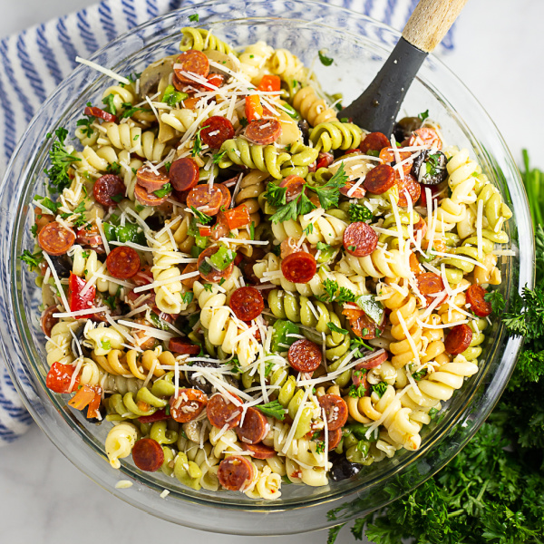 Overhead view of a large bowl of pizza pasta salad with a wooden spoon in it and a striped linen and a bunch of fresh parsley in the background.