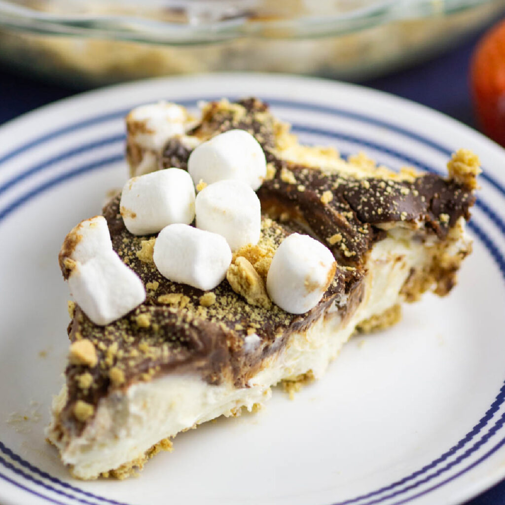 A slice of no bake s'mores cheesecake topped with mini marshmallows and graham cracker crumbs on a small plate.