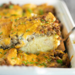 A serving of easy egg breakfast casserole topped with melted cheese and fresh chives being lifted from the remaining casserole in a white ceramic casserole dish.