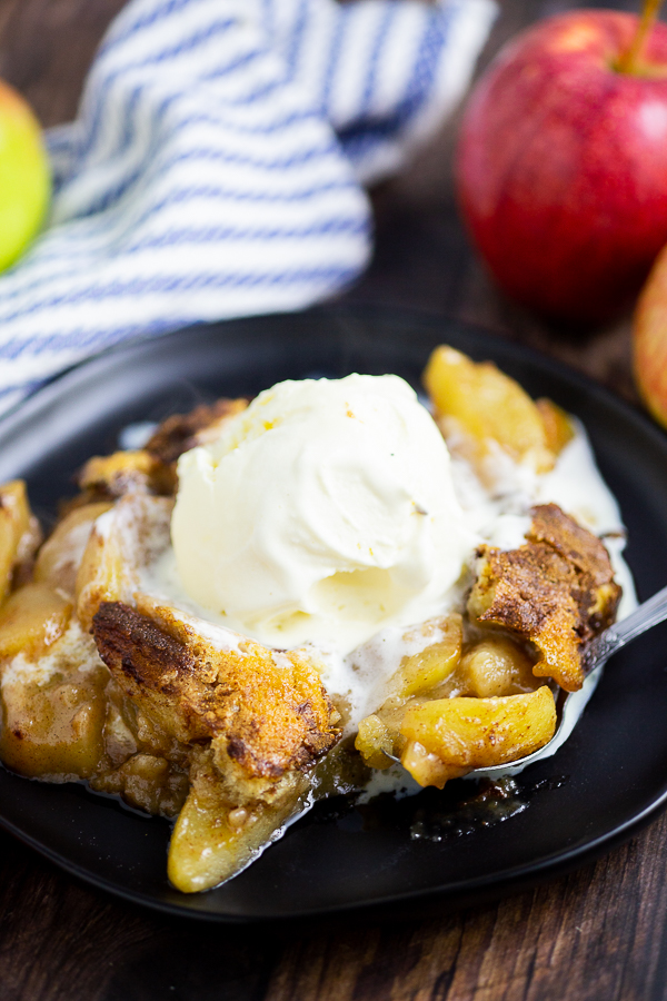 Apple Cobbler on a black plate with a scoop of ice cream on top. Linen napkin and apples are behind the plate