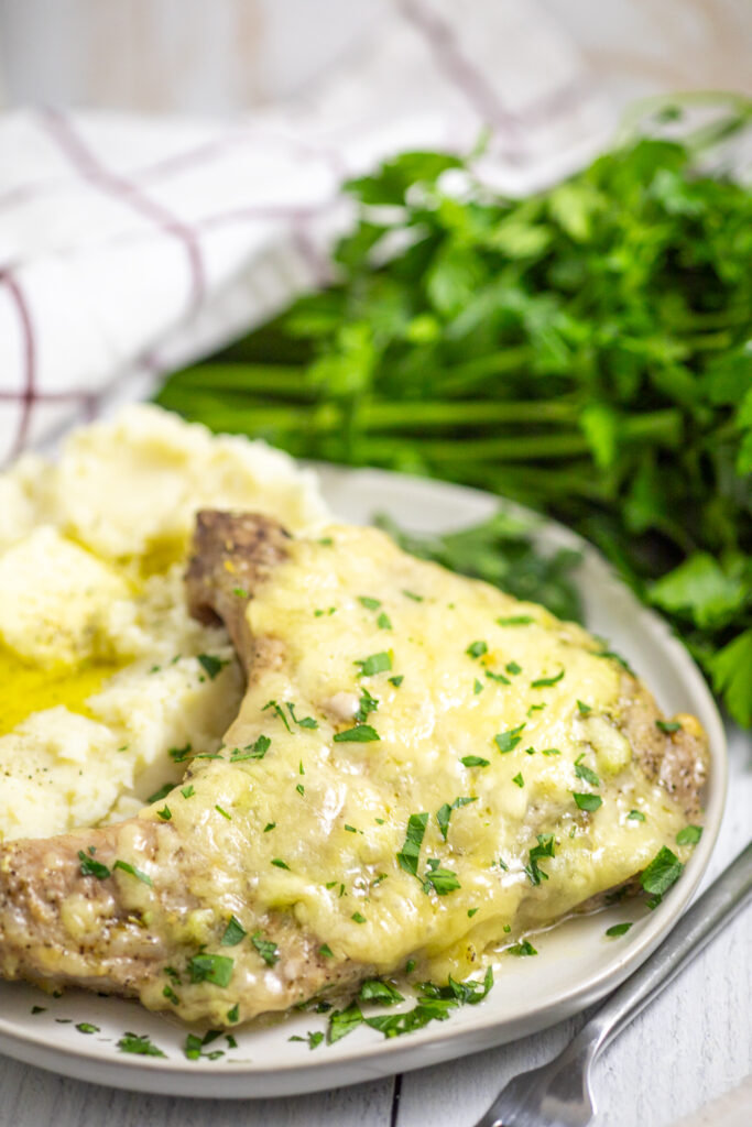 Side view of a cheesy Italian Pork Chop topped with fresh chopped parsley with a bunch of fresh parsley and a linen napkin behind.