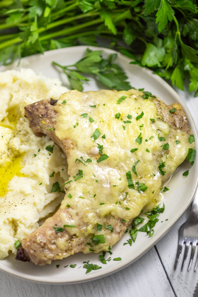 Overhead view of a cheesy Italian Pork Chop on a white plate next to buttery mashed potatoes with fresh parsley sprinkled on top with a bunch of fresh parsley behind.