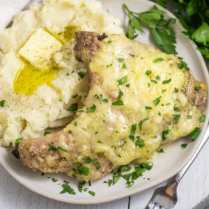 Overhead view of a cheesy Italian Pork Chop on a white plate next to buttery mashed potatoes with fresh parsley sprinkled on top.