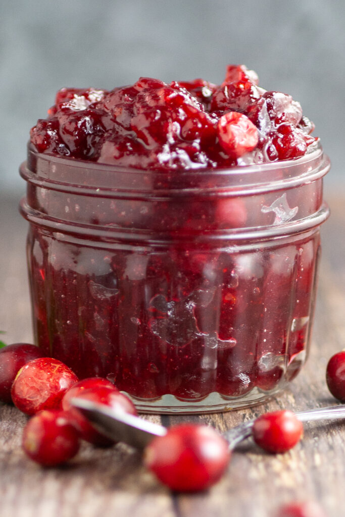 Side view of homemade cranberry sauce in a small glass jar with fresh cranberries and a spoon in front.
