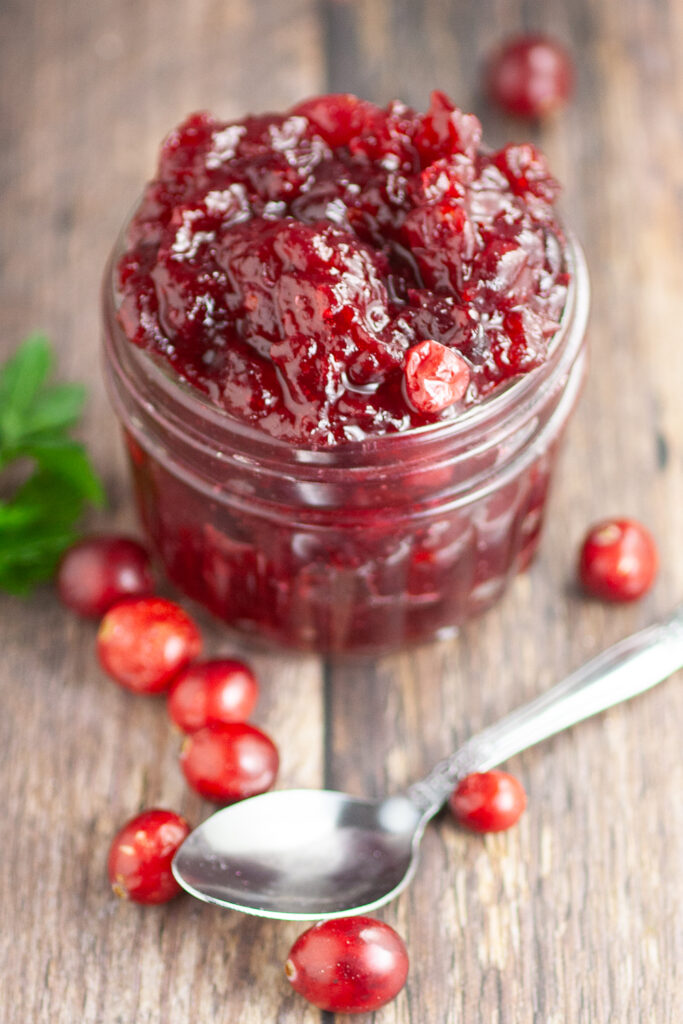 Overhead view of homemade cranberry sauce in s small jar on a rustic wood background next to fresh cranberries and a spoon.