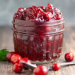 Side view of homemade cranberry sauce in a small glass jar with fresh cranberries and a spoon in front.