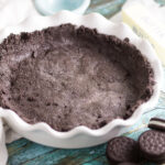 An empty Oreo crust surrounded by full chocolate sandwich cookies on a teal rustic wooden background with a linen napkin behind.