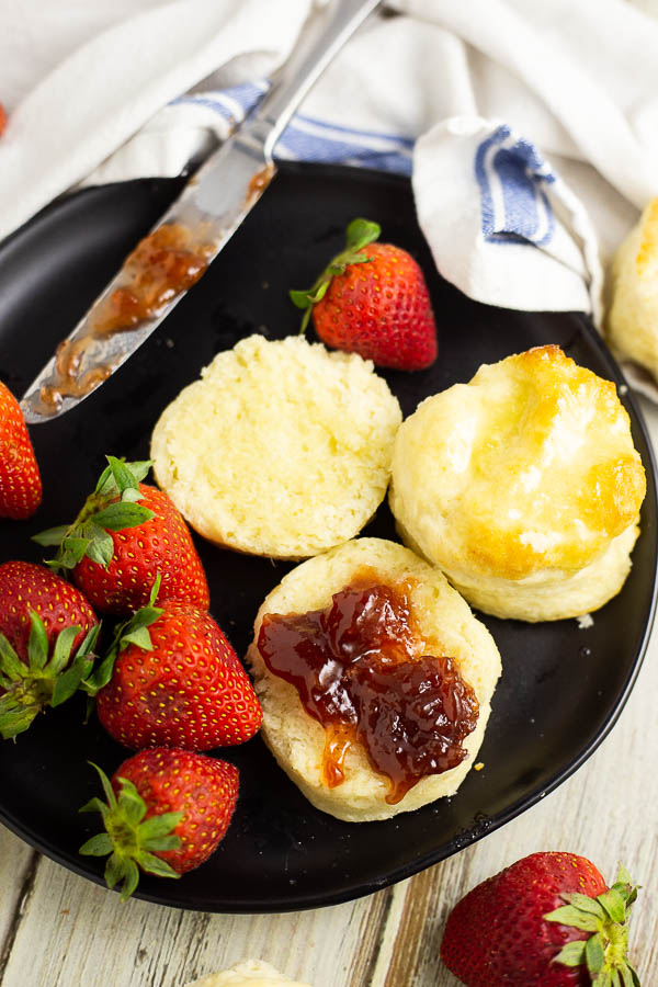 Two biscuits on a black plate plate with strawberries. One biscuit is cut open with strawberry jam spread on it
