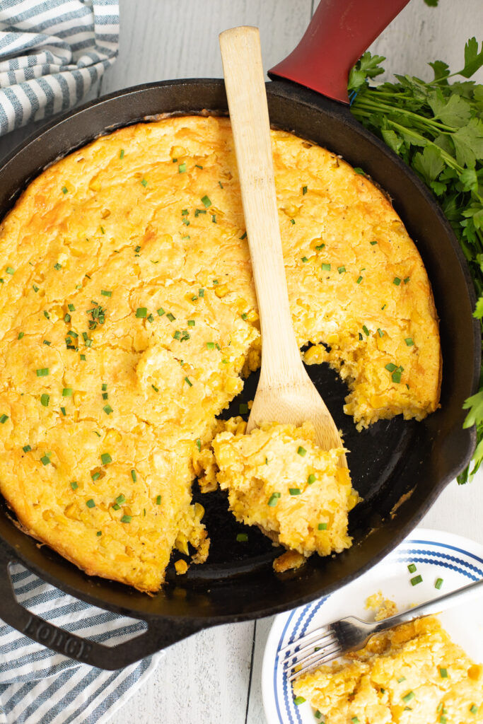 Overhead view of a wooden spoon taking a scoop of southern corn pudding from a cast iron skillet.