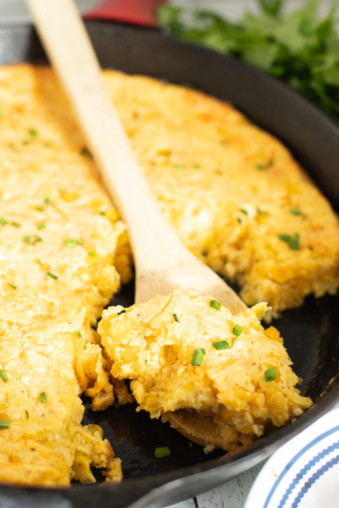 Close up view of a wooden spoon taking a scoop of southern corn pudding from a cast iron skillet.