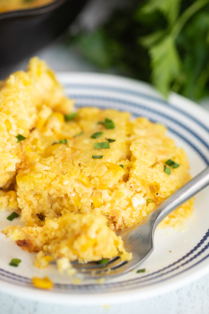 Close up view of southern corn pudding topped with fresh chives on a small white plate with a fork taking a bite off.