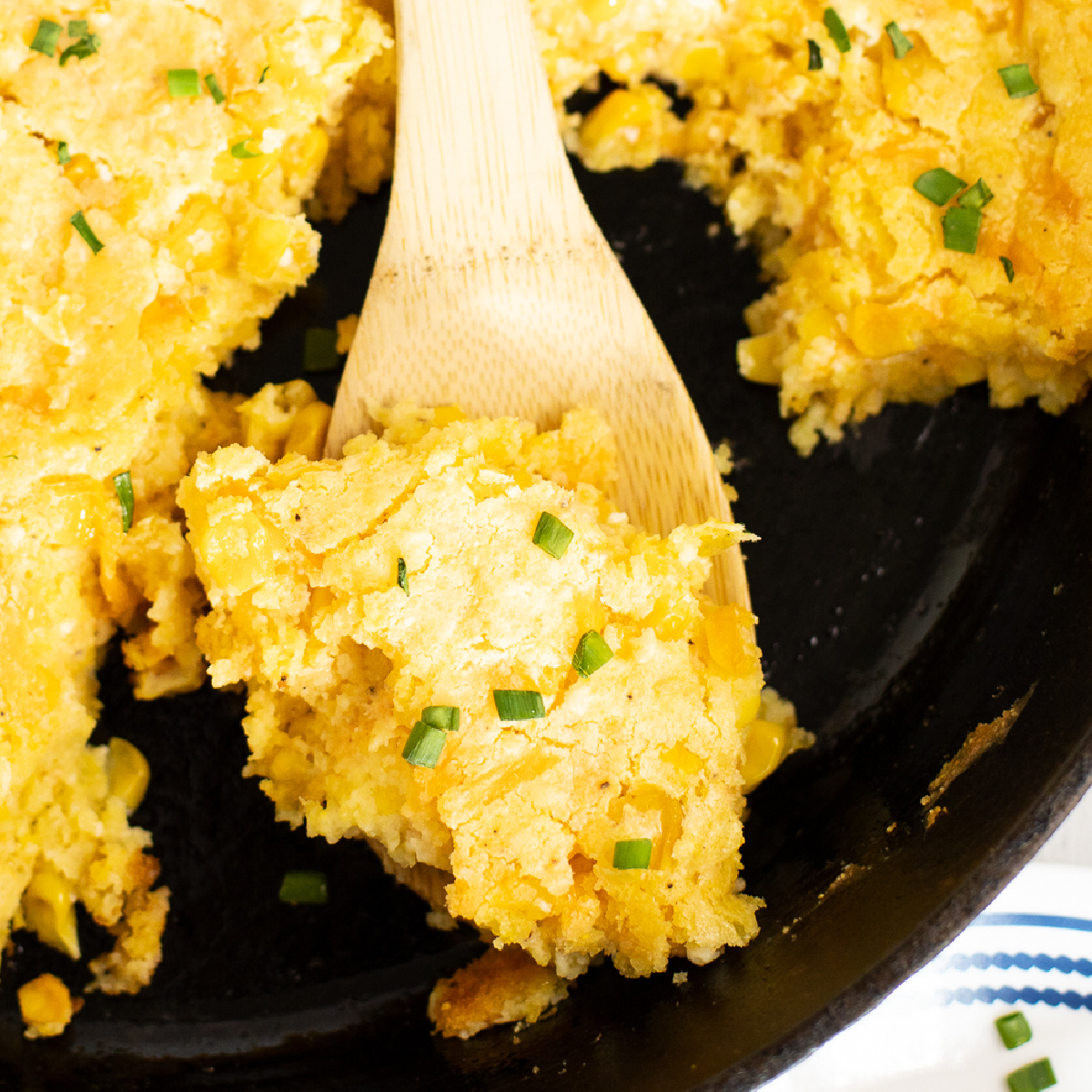 Overhead view of a wooden spoon taking a scoop of southern corn pudding from a cast iron skillet.
