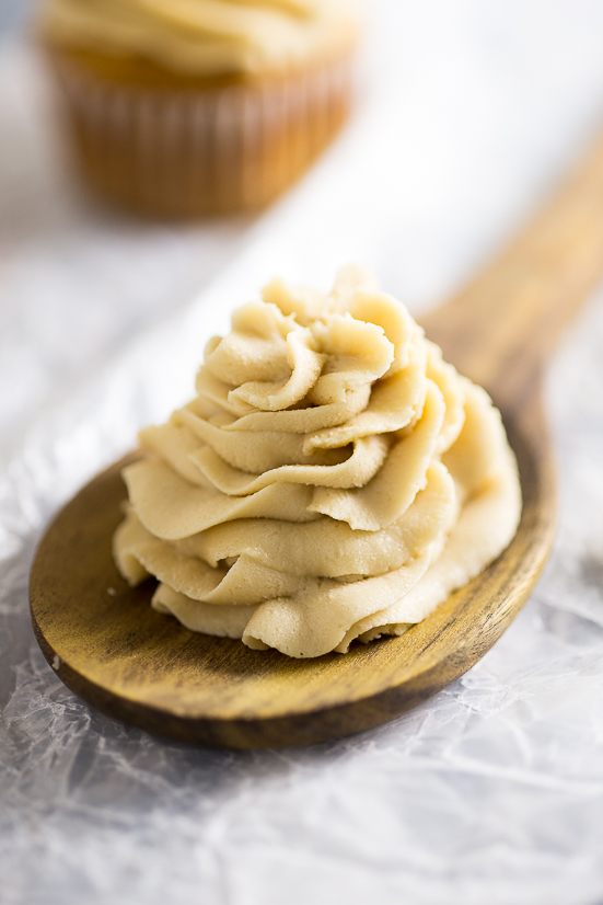 Caramel Frosting piped onto a wooden spoon on wax paper with frosted cupcakes behind