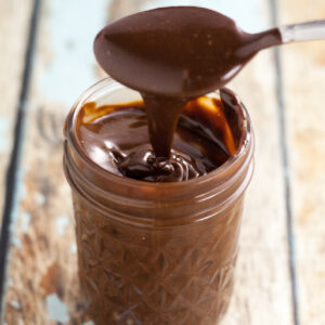 A spoon drizzling chocolate ganache into a quilted mason jar on a rustic wood background.